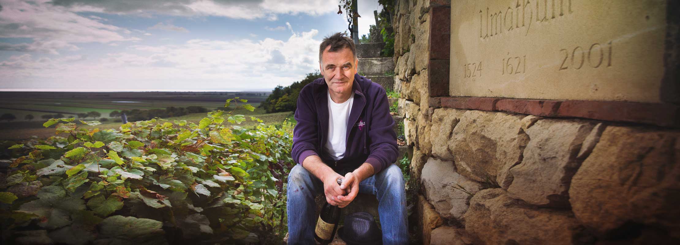 Josef Umathum sitzt im Weingarten neben einer Steinmauer mit der Inschrift „Umathum“