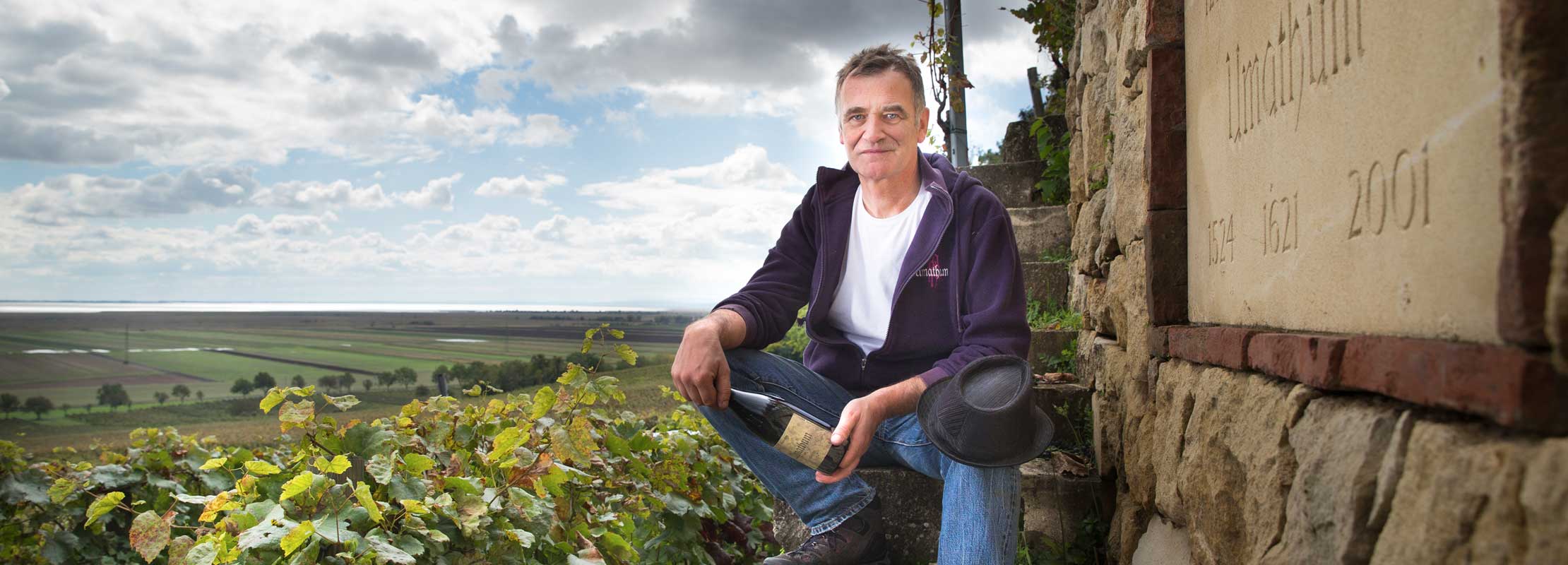 Josef Umthum sitzt neben einer Steinmauer mit einer Weinflasche in der Hand vor Landschaft mit Weingärten und Neusiedlersee im Hintergrund